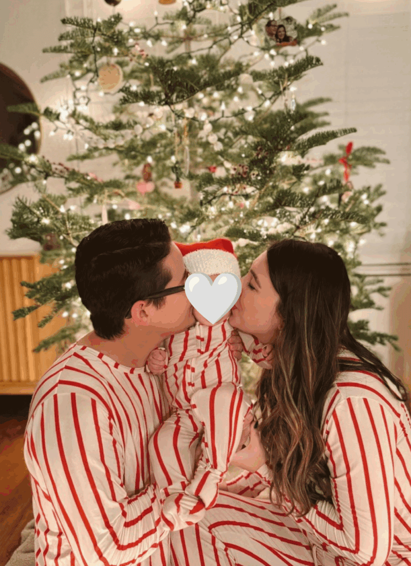 Mom and dad in matching red and white striped Christmas pajamas kissing their baby in front of a lit Christmas tree